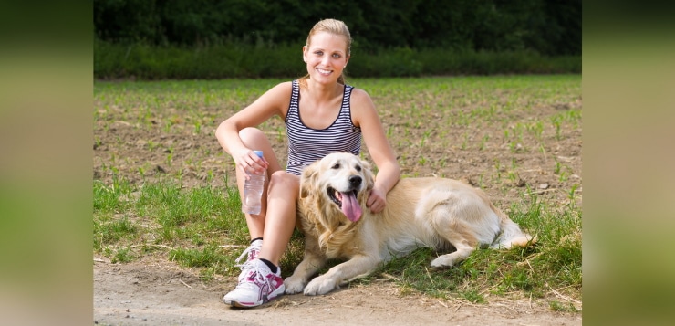 Frau hält eine Wasserflasche in der Hand und sitzt mit ihrem Golden Retriever am Wegrand, um eine Pause vom Training zu machen