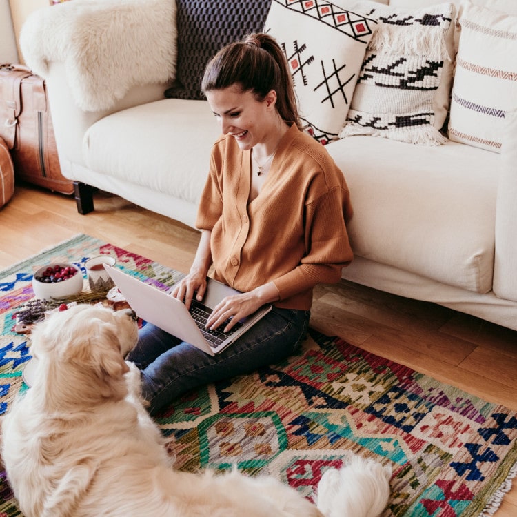 Frau sitzt auf dem Boden vor einem Sofa und hat einen Laptop auf den Beinen. Zu ihren Füßen liegt ein Golden Retriever